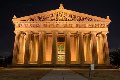 Night view of a classical Greek-style building with illuminated columns and sculptures.