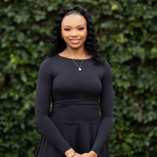 A smiling woman in a black dress stands confidently outdoors against a leafy background.