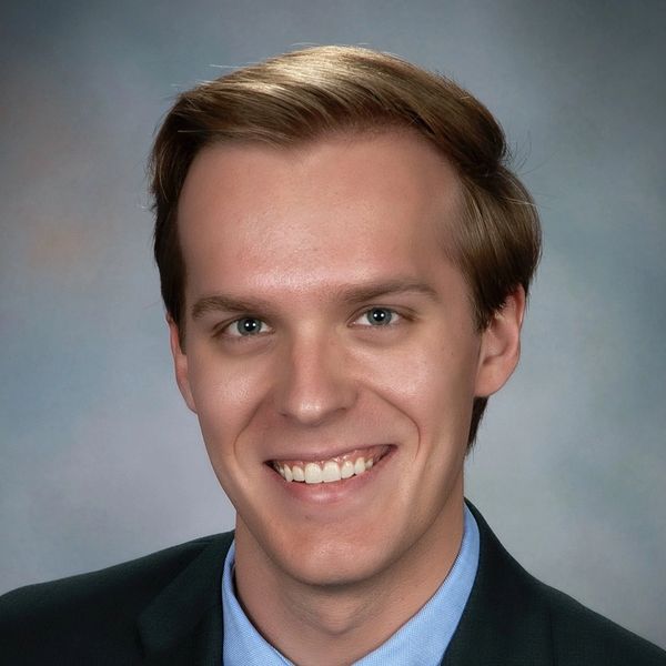Smiling young man in business attire against a soft gradient background.