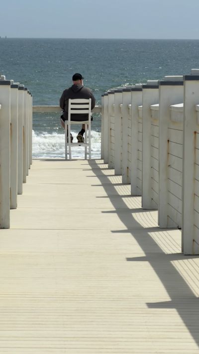 A person sits alone on a chair facing the ocean at the end of a boardwalk.