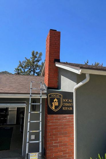 Red brick chimney with a SoCal Chimney Repair sign and a ladder beside it.