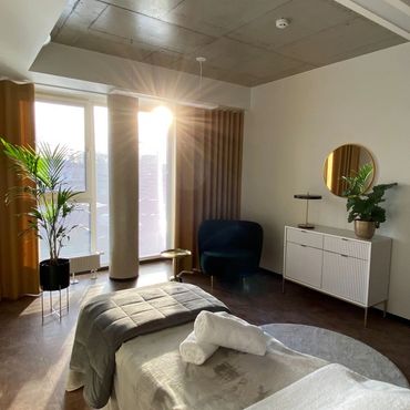 Ilukunsti hubane hoolduste ruum kliendivoodi, tugitooli ja lillega, aknast kumab päikesevalgus.