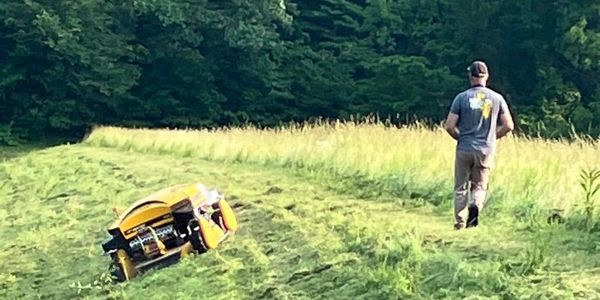 A man walks away from an overturned yellow lawn mower on a grassy hill.