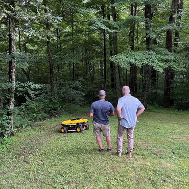 Two men operating a yellow robotic lawn mower in a lush forest clearing.
