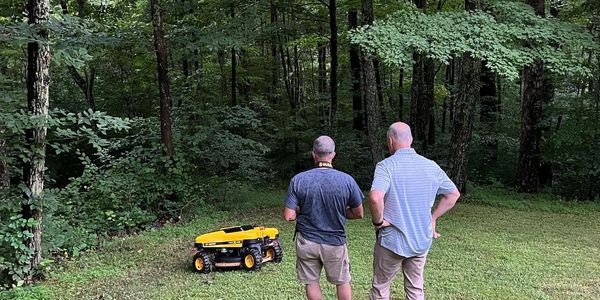 Two men operating a yellow robotic lawn mower in a lush forest clearing.
