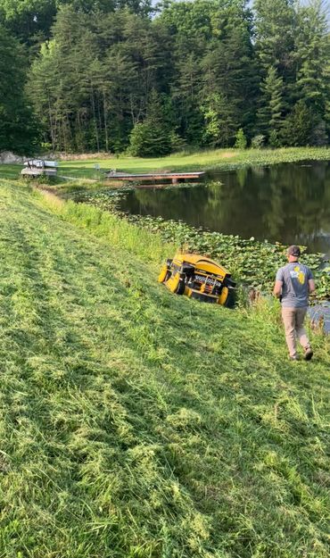 Man remotely operates a robotic mower on a steep grassy slope near a pond.