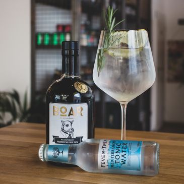 A bottle of gin, a bottle of Tonic and a Gin and Tonic garnished with Rosemary arranged on a table