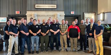 Group photo of 20 people standing indoors in front of a decorative wall and fireplace.