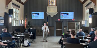 Man giving a presentation in a high-ceilinged room with attendees seated.