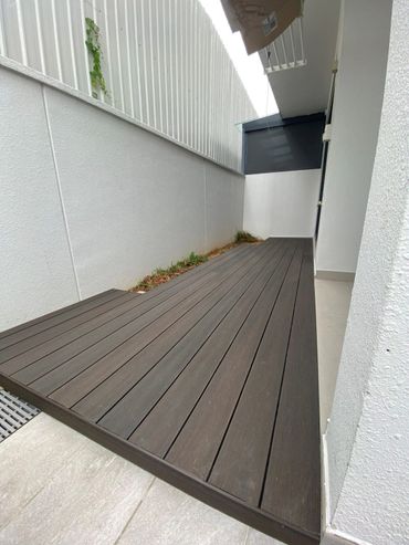 A narrow outdoor patio with dark wooden deck flooring and white walls.