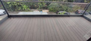 Spacious balcony with wooden deck and glass railing overlooking greenery and parked cars.