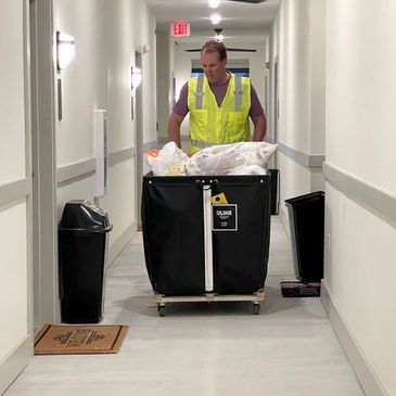 Worker pushing a large cart filled with trash bags down a hallway.
