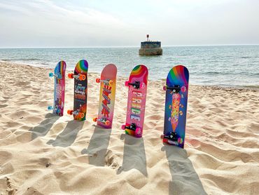 Colorful skateboards with LGBTQ+ themes stand on a sandy beach near the ocean.