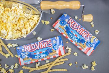 Two Red Vines Buttered Popcorn packs surrounded by popcorn and a popcorn popper.