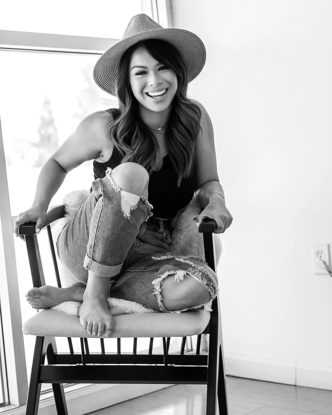 Smiling woman in a hat sitting barefoot on a chair by the window.