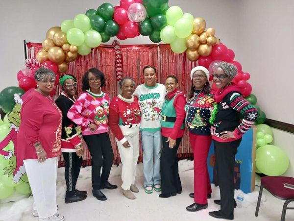 Group of women in festive Christmas sweaters posing under a balloon arch.