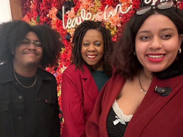 Three people smiling in front of a colorful flower wall.