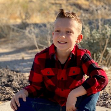 Young boy in red plaid shirt and boots sitting outdoors on concrete.