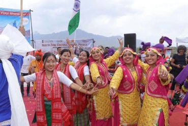 Women in traditional attire joyfully dancing at a cultural event with an Indian flag in the background.