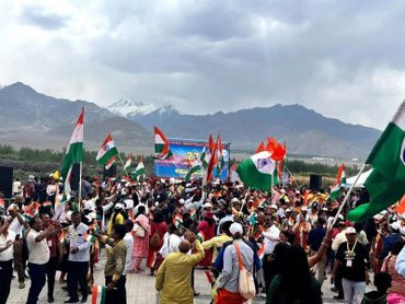 Crowd waving Indian flags with mountainous backdrop under cloudy sky.