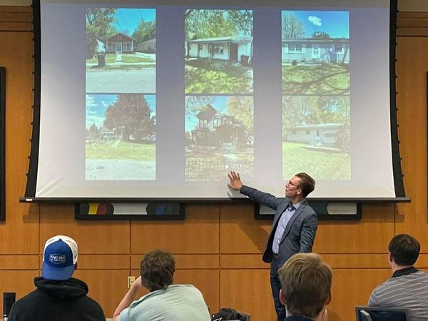 A presenter discusses house photos in a classroom setting with attentive students.