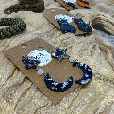 Handcrafted celestial-themed earrings displayed on fabric with braided bracelets.