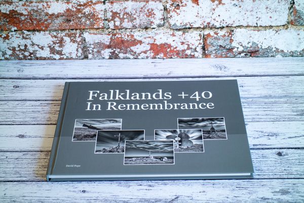 A book titled 'Falklands +40 In Remembrance' on a wooden surface with a brick backdrop.