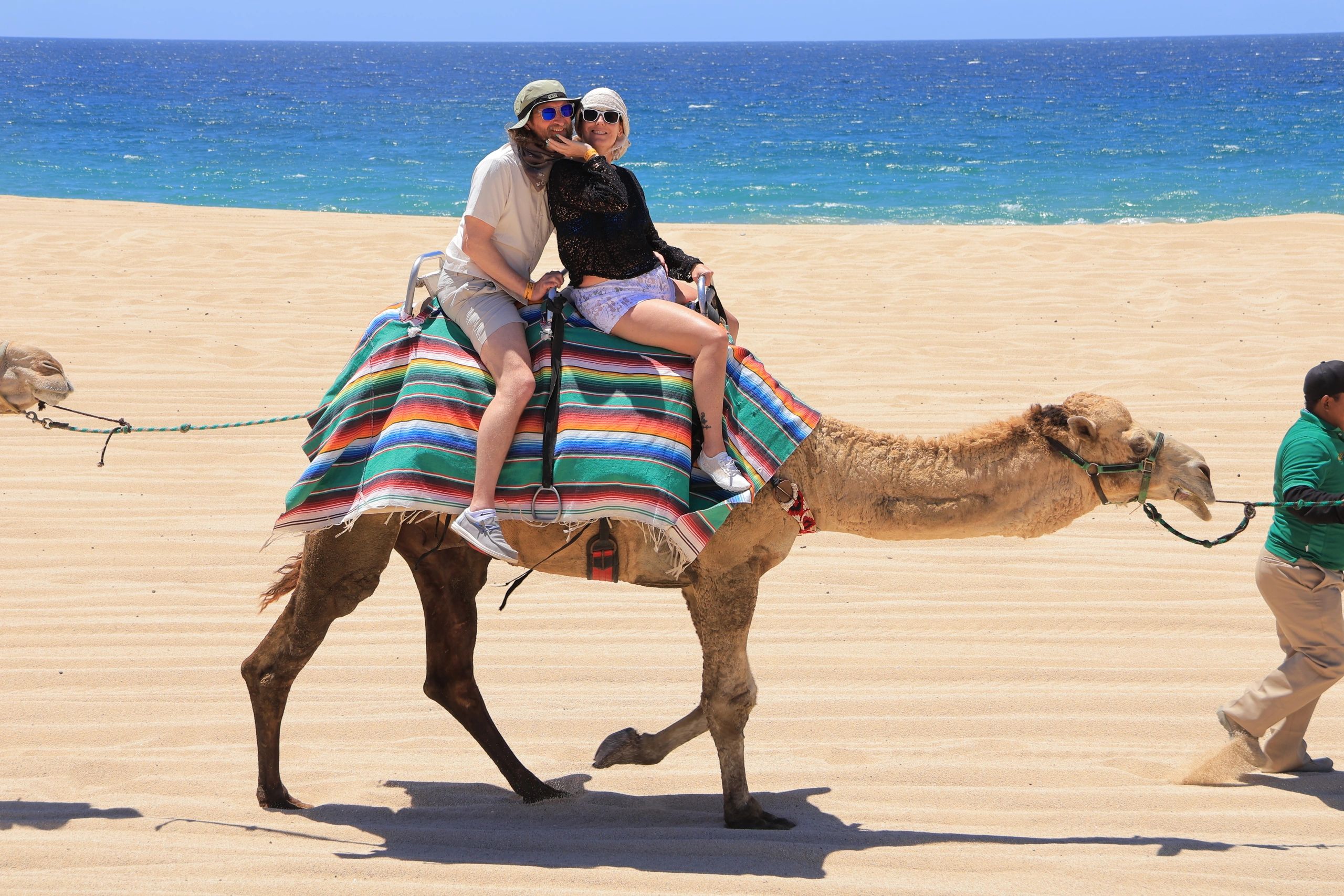 Couple enjoying a camel ride on a sunny beach with a vibrant blanket.