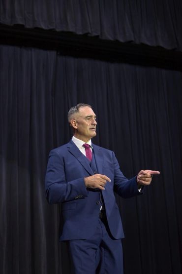 Man in blue suit pointing, standing against black curtain background.