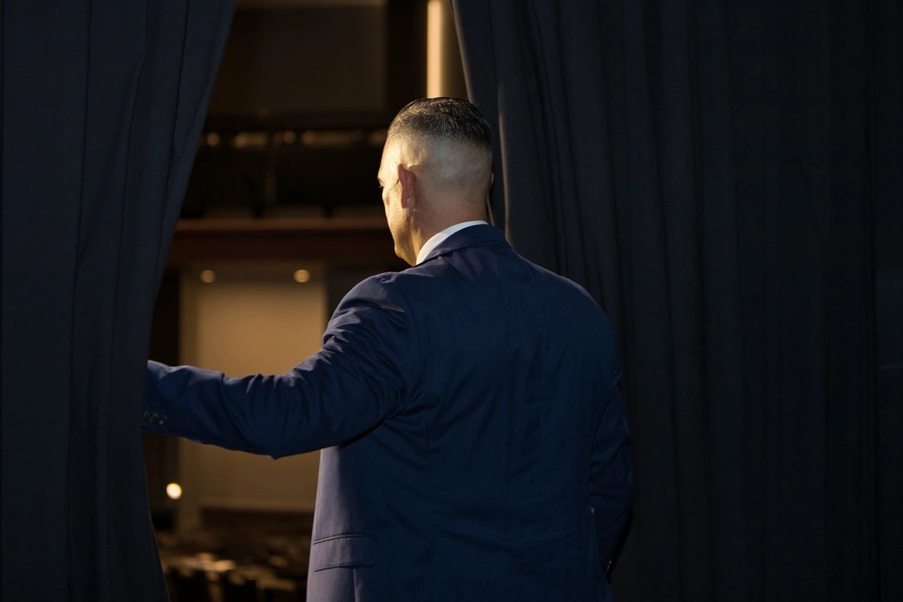 Man in a suit opening stage curtains, facing away.