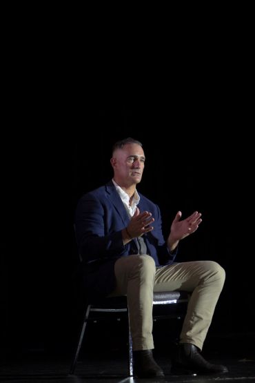 Man in blue blazer speaking expressively while seated on a chair.
