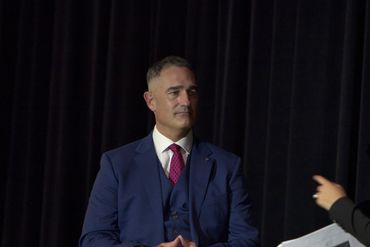 Two professionals engaged in a formal discussion on stage with dark curtains in the background.
