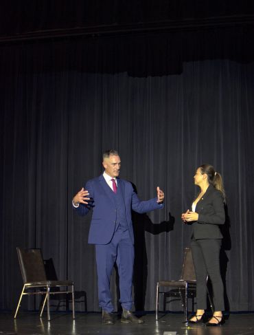 Man and woman in business attire conversing on stage with chairs behind them.