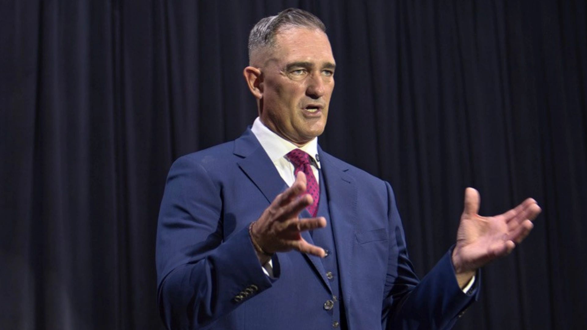 Man in blue suit speaking passionately with expressive hand gestures.