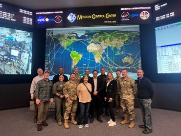 Group photo of diverse personnel at NASA Mission Control Center with global maps and mission data screens.