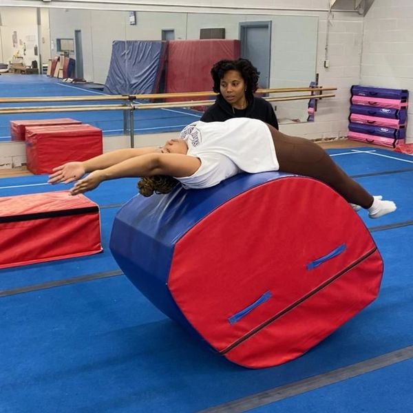Gymnast stretching on a foam barrel with coach observing.