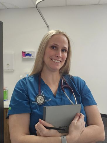 Smiling nurse in blue scrubs holding a tablet and pen in a medical office.