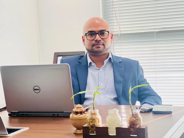 Man in a blue suit at a desk with a laptop and plants.