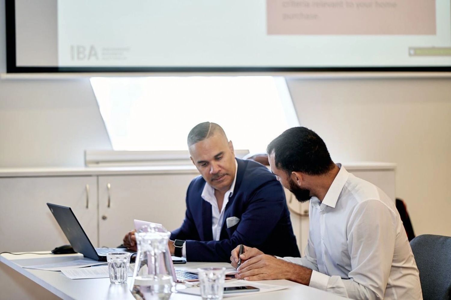 Two men in business attire discussing work at a conference table with documents and a laptop.