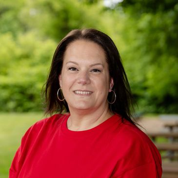 Woman in a red shirt smiling outdoors with greenery in the background.