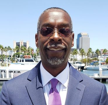 Man in sunglasses and suit smiling with a marina and cityscape in the background.