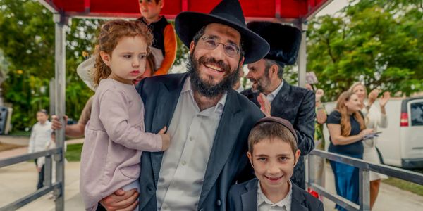 A smiling man in traditional Jewish attire holding a child, with another child beside him.