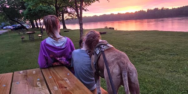 Two girls and a dog watch a colorful sunset by the river from a wooden picnic table.