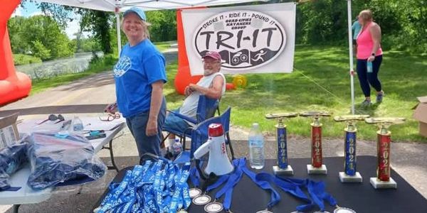 Volunteers at an outdoor event table with medals, trophies, and a banner for TRI-IT.