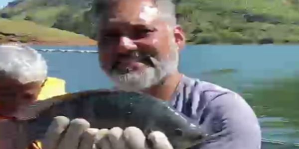 Man proudly holding a large fish by a lake with a child nearby.
