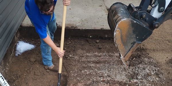 Person digging soil near a concrete slab with a shovel and excavator bucket to perform earthworks and excavation tasks such as preparing a trench for a new power line.
