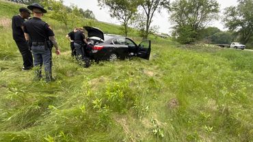 Police officers inspecting a black car in a grassy area after an incident.