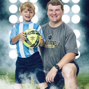 A young boy and man smiling, holding a soccer ball with bright lights behind.