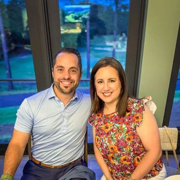 Smiling couple sitting indoors near large windows with a scenic view outside.