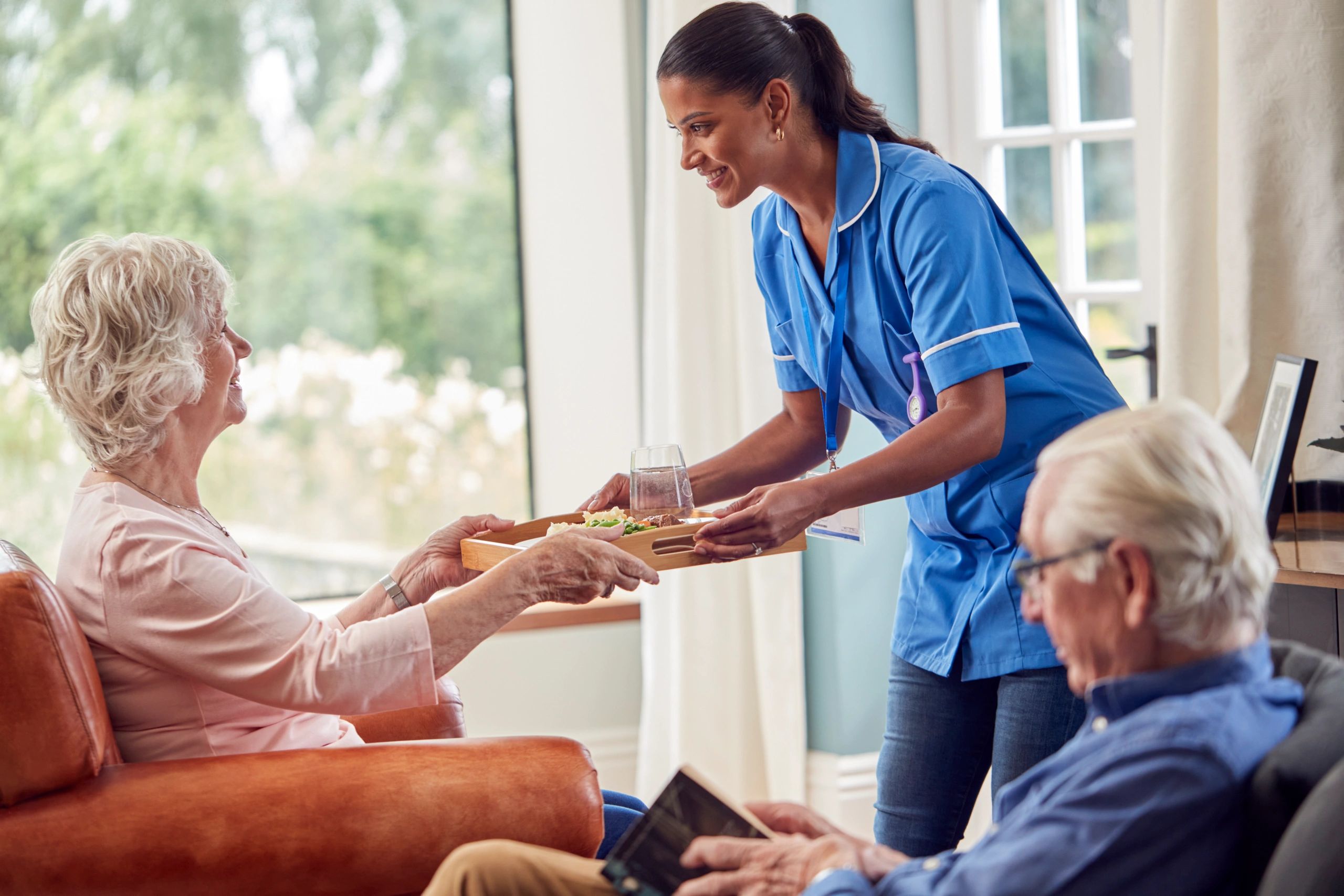 A caregiver in blue scrubs is assisting an elderly person in a seated position